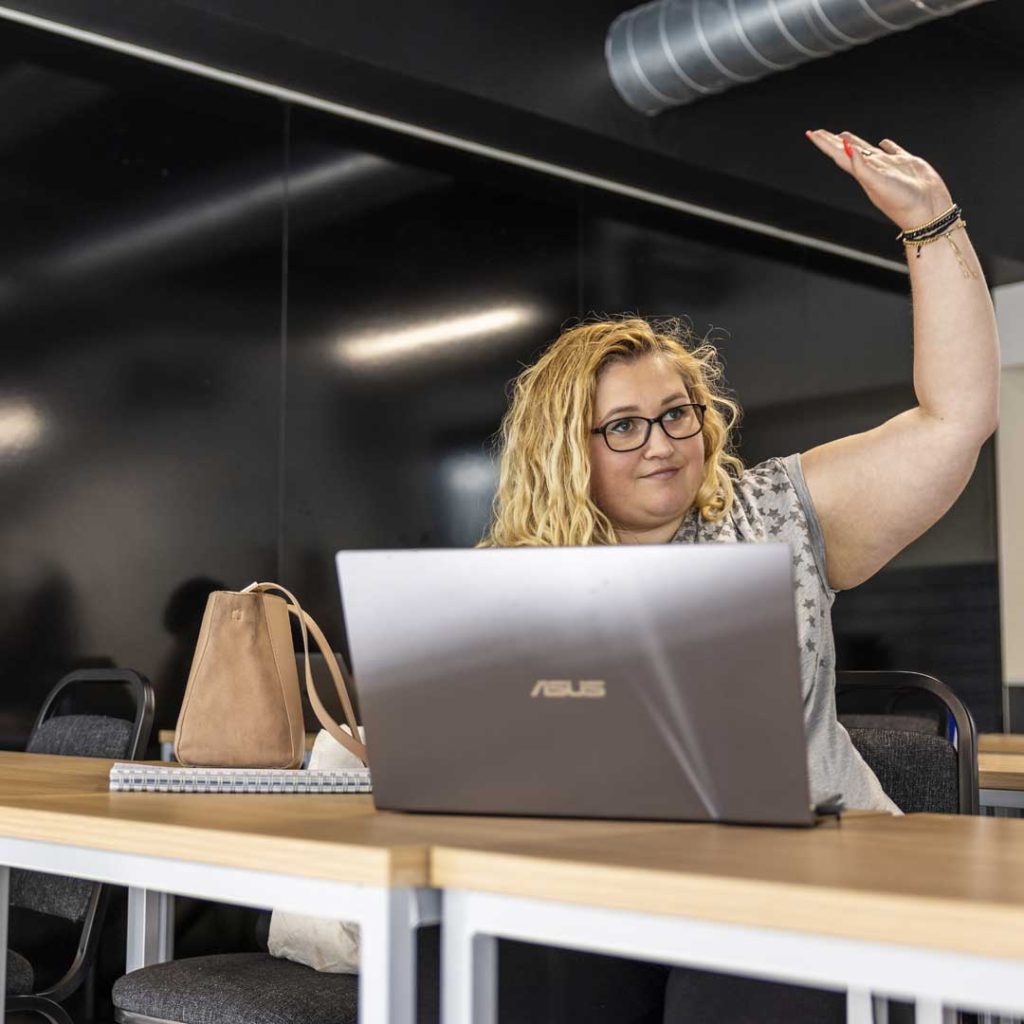 female student sat behind a laptop with her hand up in class